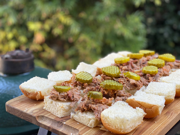 Platter of open pulled pork sandwiches topped with pickles on white buns outdoor next to a smoker