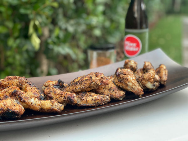 Plate of grilled seasoned chicken wings on an outdoor table next to soda bottle and glass of beer